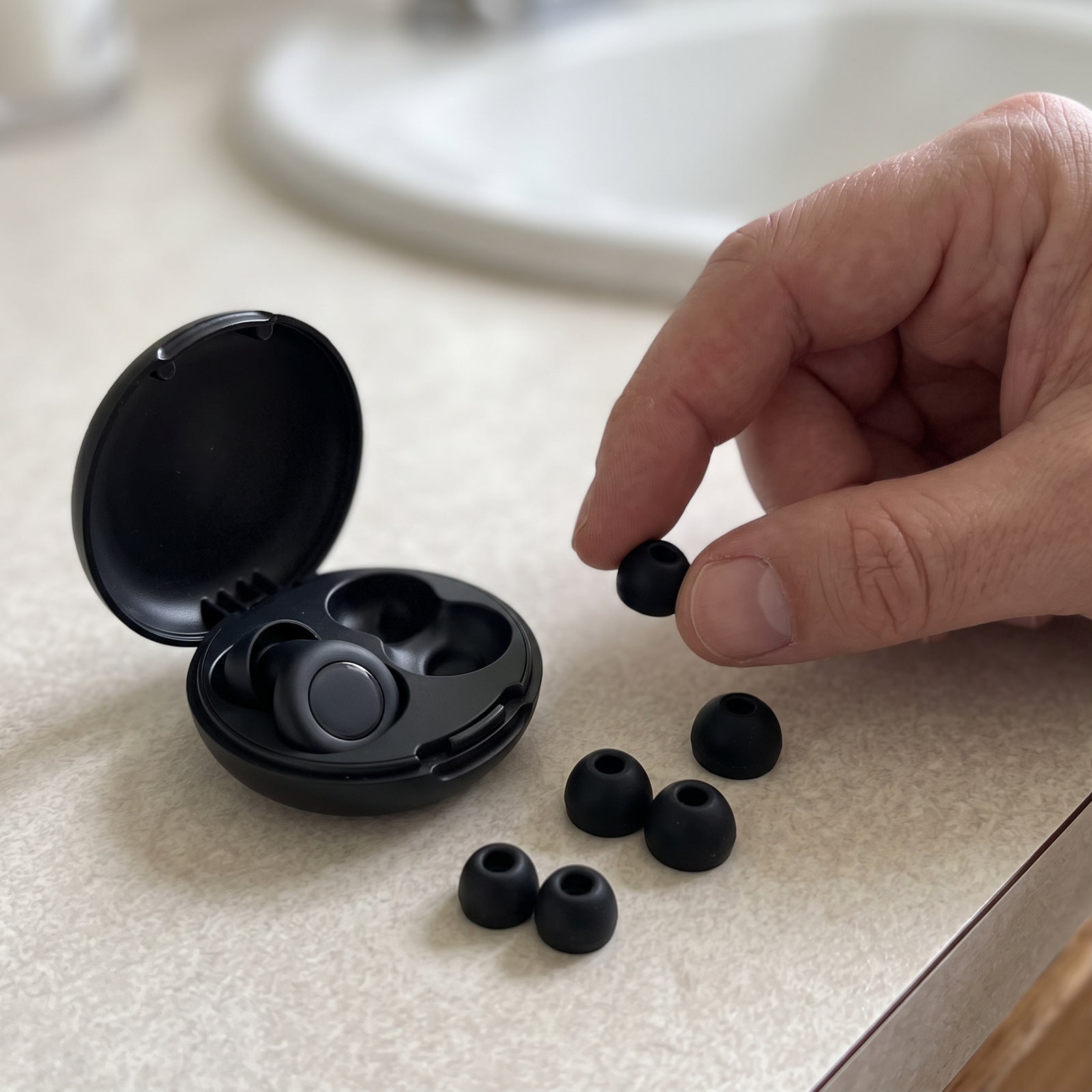 Hand choosing a silicone tip beside Black earplugs and an open round case