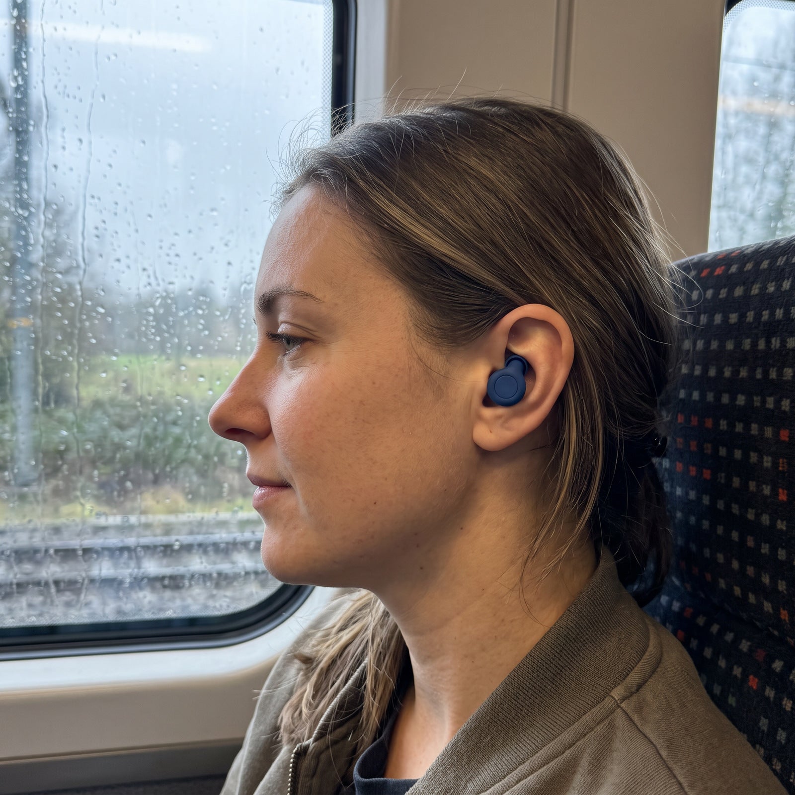 Woman wearing Blue Stillday earplugs beside a rain-streaked train window